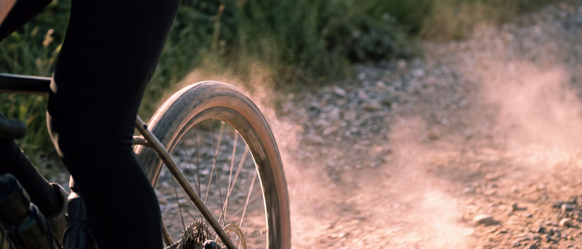 Wiel en been van een fietser op een droge onverharde weg, met stof dat opwaait bij het passeren